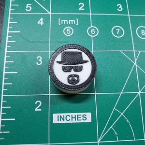 Puede incluir: Un objeto redondo, blanco y negro, con un diseño de sombrero, gafas de sol y barba. La palabra "INCHES" está impresa en la superficie verde. El objeto tiene aproximadamente 2,5 cm de diámetro.