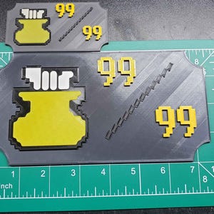 Peut inclure: Un objet de jeu imprimé en 3D de couleur grise et jaune avec une fiole de potion jaune et le nombre 99 imprimé dessus. L'objet est sur une surface grise avec une règle en arrière-plan.