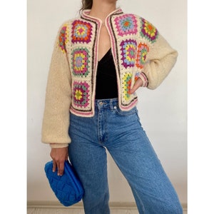 May include: A cream-colored crocheted cardigan with a colorful granny square pattern on the front. The cardigan has long sleeves and a button closure.