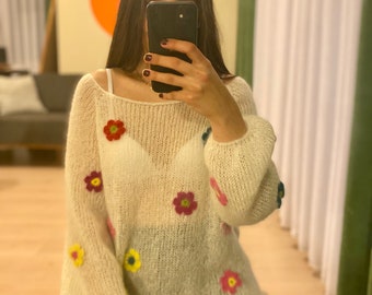 Puede incluir: Suéter de punto abierto color crema adornado con flores bordadas de colores. El suéter tiene un escote amplio y mangas largas abullonadas. Los adornos florales incluyen detalles en rojo, amarillo, rosa y azul, creando una estética artesanal y caprichosa.