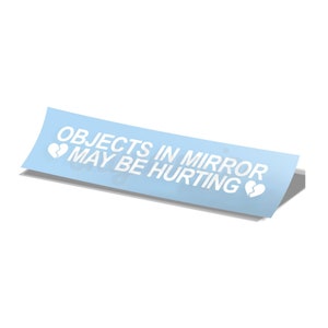 Puede incluir: Una pegatina azul claro con el texto "OBJECTS IN MIRROR MAY BE HURTING" en texto blanco con dos símbolos de corazón roto a cada lado del texto.