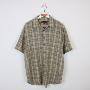 May include: A beige and brown plaid short-sleeved button-down shirt with a pocket on the left side. The shirt has a dark brown button closure and a label that reads "Pacific Trail" on the front.