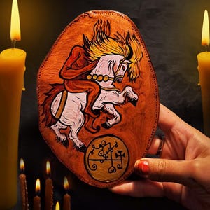 Könnte beinhalten: Handbemalte, ovale Holzplakette mit einem Reiter auf einem weißen Pferd mit gelber Mähne. Der Reiter ist rot dargestellt, und unten befindet sich ein goldenes Siegel mit Symbolen. Der Hintergrund ist warmbraun, die Szene wird von Kerzen beleuchtet.