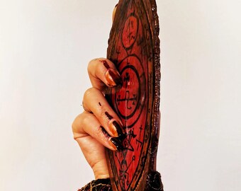 May include: A wooden, shield-shaped object with a red background and black symbols. The edges are dark brown, and the surface has a textured appearance. The design includes circular and cross-like patterns. The object is held against a white background.