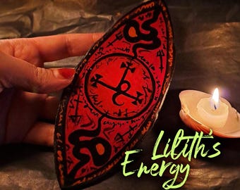 May include: A red, shield-shaped item with black symbols and text, held by a hand. The text "Lilith's Energy" is written in green. A lit candle in a seashell is in the background.