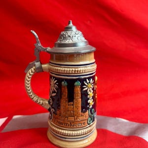 May include: A ceramic beer stein with a metal hinged lid. The stein features a detailed illustration of a building with the text "Frauentürme". The handle is beige and the body is blue, beige, and brown.