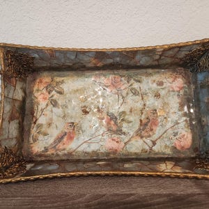 May include: A rectangular serving tray featuring a mosaic-style border in shades of blue and brown with gold-toned metal handles and corner accents. The centre panel displays a printed illustration of birds perched on branches among pink roses.