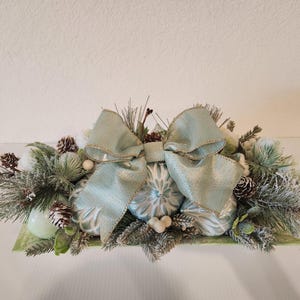 Puede incluir: Centro de mesa navideño festivo con adornos azul claro, piñas y vegetación artificial. Un gran lazo azul claro brillante con ribete dorado adorna el conjunto. Ideal para la decoración navideña.