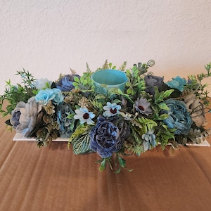 Puede incluir: Un centro de mesa floral azul y verde con un portavelas turquesa. El centro de mesa está sobre una base rectangular blanca.