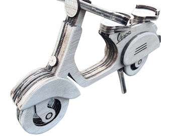 May include: A white wooden model of a Vespa scooter. The scooter has a black seat and handlebars, and the word "Vespa" is printed on the side.