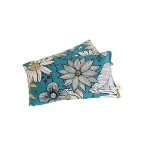 Pode incluir: Duas almofadas retangulares de tecido azul-petróleo com um padrão floral. O padrão apresenta grandes flores brancas e creme com contornos pretos e detalhes amarelos. As almofadas são provavelmente decorativas ou para conforto.