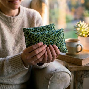 Pode incluir: Dois pacotes de calor retangulares verdes com um padrão de leopardo preto e azul. Os pacotes são segurados em frente a uma pessoa vestindo um suéter bege. O fundo inclui uma janela, uma cadeira e uma mesa lateral com uma xícara de café.