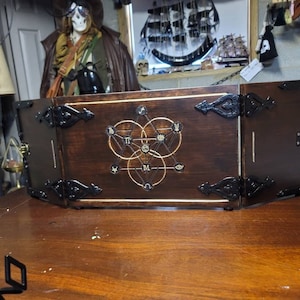 Puede incluir: Una caja de madera con tapa con bisagras y detalles en metal negro. La tapa presenta un diseño geométrico con un círculo, una estrella y un triángulo. La caja está cerrada.