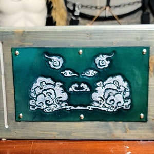 May include: A three-panel wooden box with a green background and white cloud designs. The box is closed and has a rustic finish.