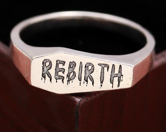 May include: A silver ring with the word "REBIRTH" engraved on it. The letters are in a dripping font style.