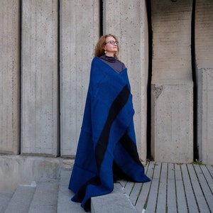 Op de afbeelding: Een vrouw met een bril staat voor een betonnen muur, gewikkeld in een blauwe en zwarte deken.