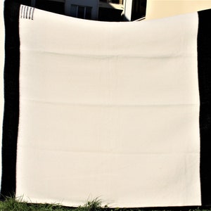 Puede incluir: Una manta de lana blanca con bordes negros. La manta est&aacute; doblada y sostenida por dos manos.