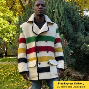 Op de afbeelding: Een crèmekleurige, dubbelbreasted jas met horizontale strepen in groen, rood, geel en zwart. De jas heeft een brede kraag, twee voorzakken en zwarte bies. De persoon draagt een blauwe spijkerbroek.