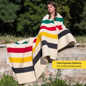 Puede incluir: Una manta color crema con rayas horizontales en verde, rojo, amarillo y negro. La manta está sobre una persona sentada al aire libre. El texto "Free Express Delivery US Tariffs & Duties paid." es visible.