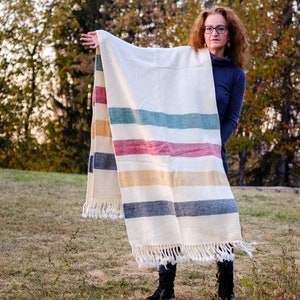 Op de afbeelding: Een vrouw houdt een witte deken vast met meerkleurige strepen. De deken heeft een franje rand en is gemaakt van een zachte, geweven stof.