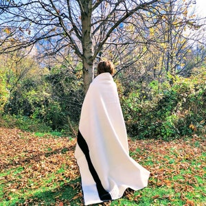 Puede incluir: Una persona de pie en un parque usando una manta blanca con una franja negra. La manta est&aacute; drapeada sobre los hombros de la persona y le cubre la cabeza. La persona est&aacute; de pie frente a un &aacute;rbol con corteza marr&oacute;n y hojas verdes. El suelo est&aacute; cubierto de hojas ca&iacute;das.