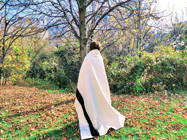 Puede incluir: Una persona de pie en un parque usando una manta blanca con una franja negra. La manta est&aacute; drapeada sobre los hombros de la persona y le cubre la cabeza. La persona est&aacute; de pie frente a un &aacute;rbol con corteza marr&oacute;n y hojas verdes. El suelo est&aacute; cubierto de hojas ca&iacute;das.