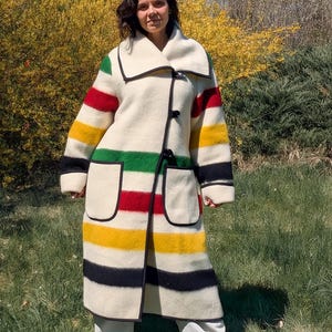 May include: A long, cream-colored coat with horizontal stripes in red, green, yellow, and black. The coat has a large collar, black trim, and two front pockets. It is fastened with three black buttons.