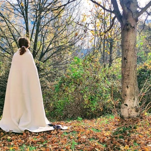 Puede incluir: Una persona con una manta o chal blanco se encuentra en un entorno boscoso con hojas ca&iacute;das en el suelo. La persona est&aacute; de espaldas a la c&aacute;mara y mirando los &aacute;rboles en la distancia.