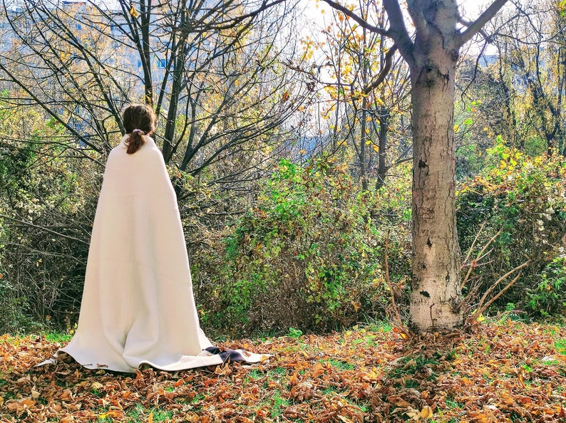 Puede incluir: Una persona con una manta o chal blanco se encuentra en un entorno boscoso con hojas ca&iacute;das en el suelo. La persona est&aacute; de espaldas a la c&aacute;mara y mirando los &aacute;rboles en la distancia.