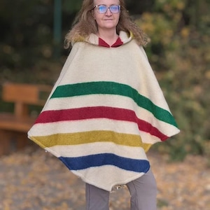 May include: Cream-colored poncho with horizontal stripes in green, red, yellow, and blue. The hooded poncho is made of a thick, woven material. The hood has a red lining. The person is wearing glasses and gray pants.