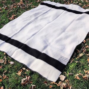 Puede incluir: Una manta de lana blanca con rayas negras. La manta est&aacute; doblada y puesta sobre un lecho de hierba verde con hojas marrones.