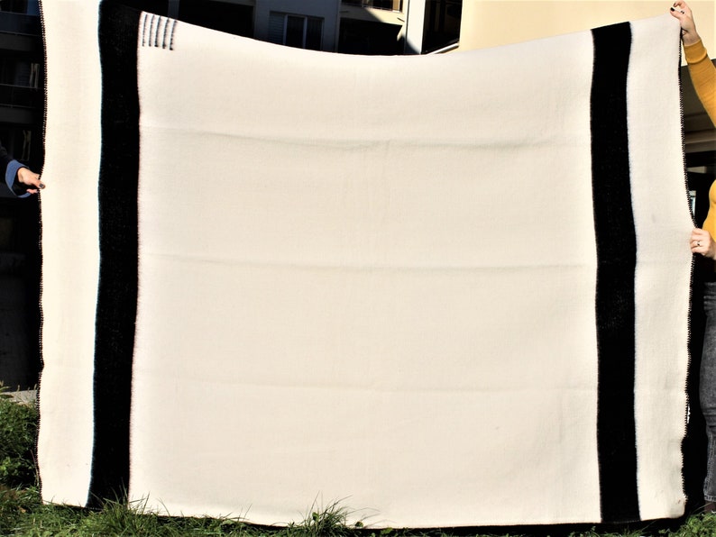 Puede incluir: Una manta de lana blanca con bordes negros. La manta est&aacute; doblada y sostenida por dos manos.