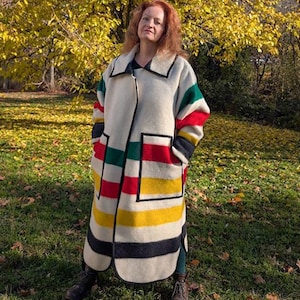 May include: A long, cream-coloured coat with a colourful striped design. The coat features stripes of red, green, yellow, and black. It has a large collar and two front pockets. The person in the image is wearing the coat outdoors.