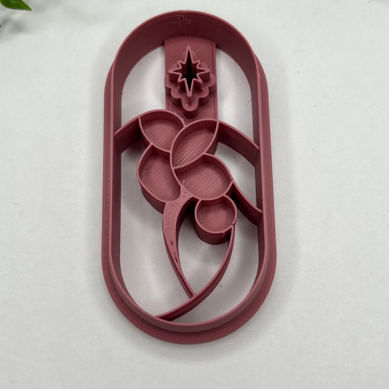 May include: A red plastic cookie cutter shaped like a stylized flower with a star in the center. The cutter is designed to create a decorative cookie shape.