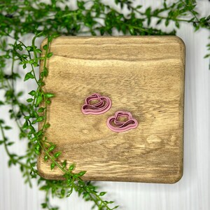 May include: Two pink plastic cookie cutters shaped like cowboy hats on a wooden surface.