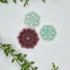 May include: Three cookie cutters in the shape of a flower. The cutters are made of metal and have a white background with a green design. The largest cutter is in the center and is a darker shade of pink.