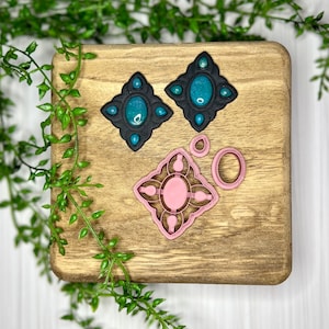 May include: A set of two black and teal resin earrings with a geometric design. The earrings are displayed on a wooden surface with a pink plastic mold for making earrings.