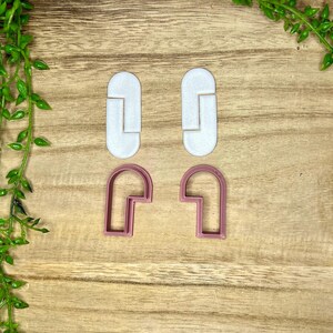 May include: Two sets of cookie cutters, one set is white and the other is pink. The white cutters are shaped like a rounded rectangle with a smaller rectangle cut out of the center. The pink cutters are shaped like a rounded rectangle with a smaller rectangle cut out of the center and a smaller rectangle cut out of the top.