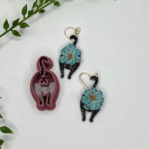 May include: Two pairs of black cat earrings with blue flower accents. One pair is a polymer clay earring and the other is a metal earring.