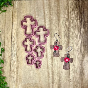 May include: A collection of pink cross-shaped cookie cutters in various sizes, alongside two wooden cross earrings with red flower accents. The items are arranged on a light brown wooden surface.