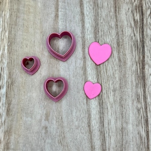 May include: Four heart-shaped cookie cutters in different sizes, two pink hearts, and one pink heart-shaped cookie cutter on a wooden surface.