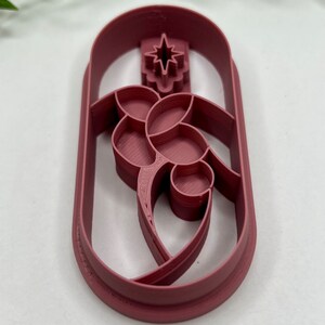 May include: A burgundy plastic cookie cutter shaped like a Christmas tree with a star on top. The tree is decorated with a stylized floral design.