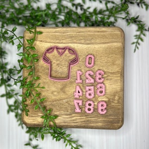 May include: Pink plastic cookie cutters in the shape of a football jersey and the numbers 0-9. The cutters are on a wooden surface.
