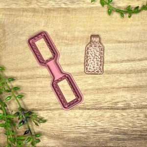 Puede incluir: Dos cortadores de galletas sobre una superficie de madera. Uno es una forma de botella rectangular rosa con un asa, y el otro es una forma de botella más pequeña y texturizada. El fondo es una veta de madera marrón claro.