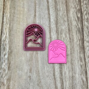 May include: Two pink plastic cookie cutters shaped like an arched window with a sun and mountains inside. The cutter on the left is a cutout design, while the cutter on the right is a solid design.