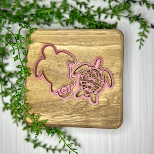 May include: Two pink plastic turtle cookie cutters on a wooden surface. The turtle on the left has a smooth shell, while the turtle on the right has a textured shell.