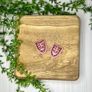 May include: Two pink plastic cookie cutters shaped like theater masks. One mask is smiling and the other is frowning.