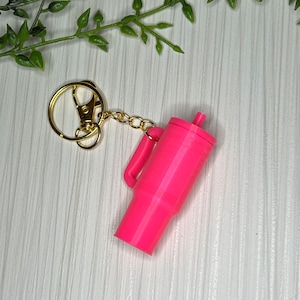 Puede incluir: Un llavero rosa con forma de vaso con una cadena y un anillo dorados.
