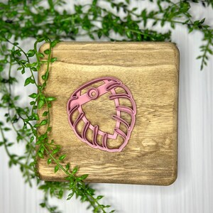 May include: A pink plastic cookie cutter in the shape of a tropical leaf, resting on a wooden cutting board. The cutter has a detailed design with multiple curved lines and a circular element. Green foliage surrounds the board.