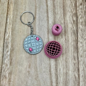 May include: A silver-coloured keychain with a waffle pattern and pink accents. The keychain is attached to a silver key ring. There are two smaller pink waffle-shaped pieces next to the keychain.
